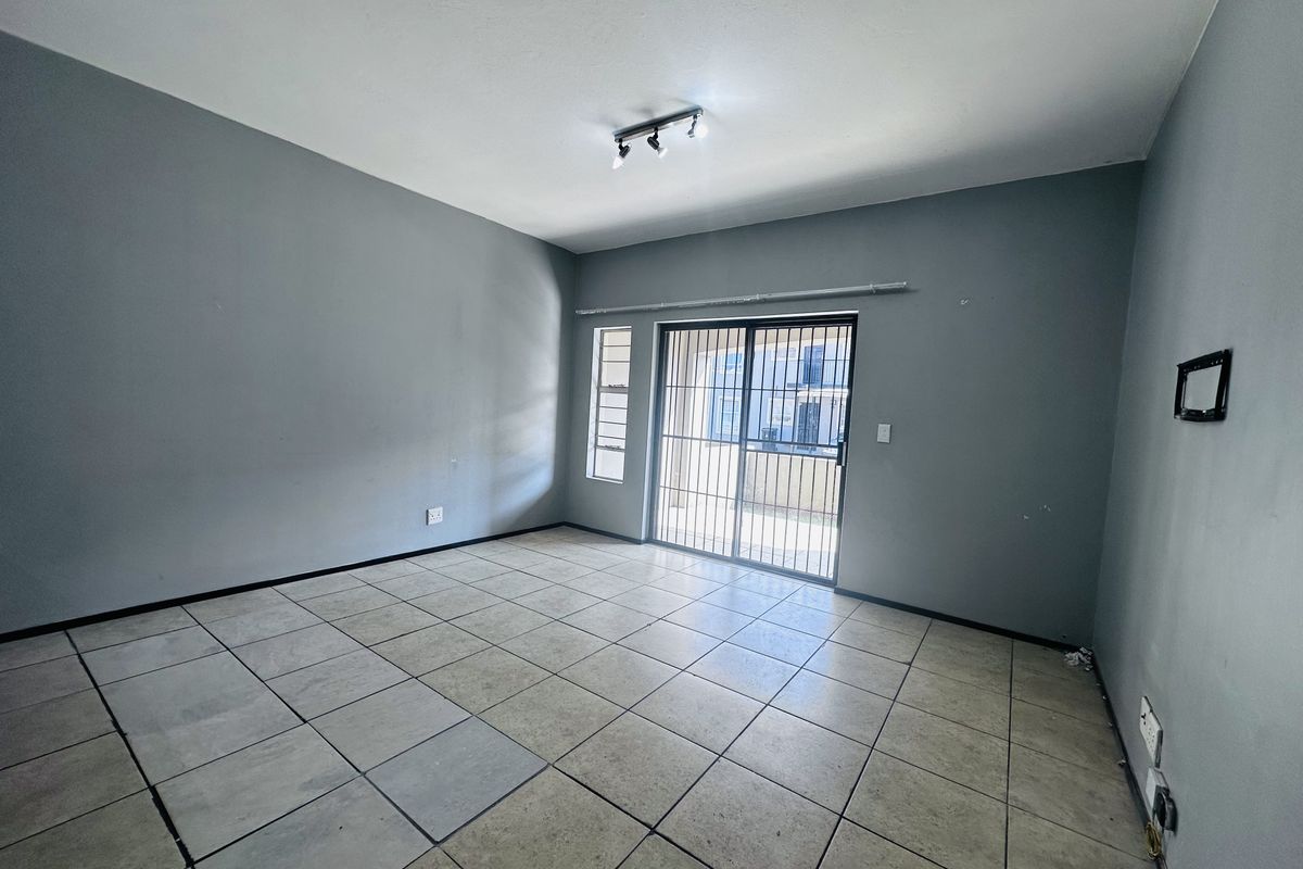 An empty, open-plan living area featuring neutral grey walls, tiled flooring, and a sliding glass door with a security gate leading to the exterior.