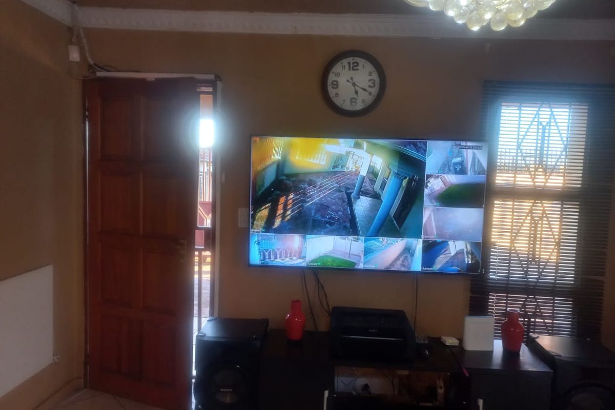 A living room area featuring a wall-mounted television displaying security camera feeds, a wooden door, and window blinds.