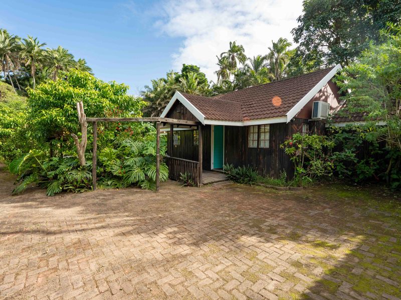 A charming timber-clad cottage featuring a tiled roof, a small timber deck entrance, and a spacious paved driveway surrounded by lush garden greenery.