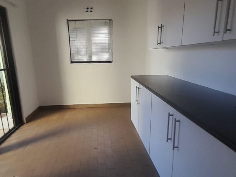 A clean, empty scullery or utility area featuring built-in white cabinetry, dark countertops, and direct access to an outdoor patio through a sliding glass door.