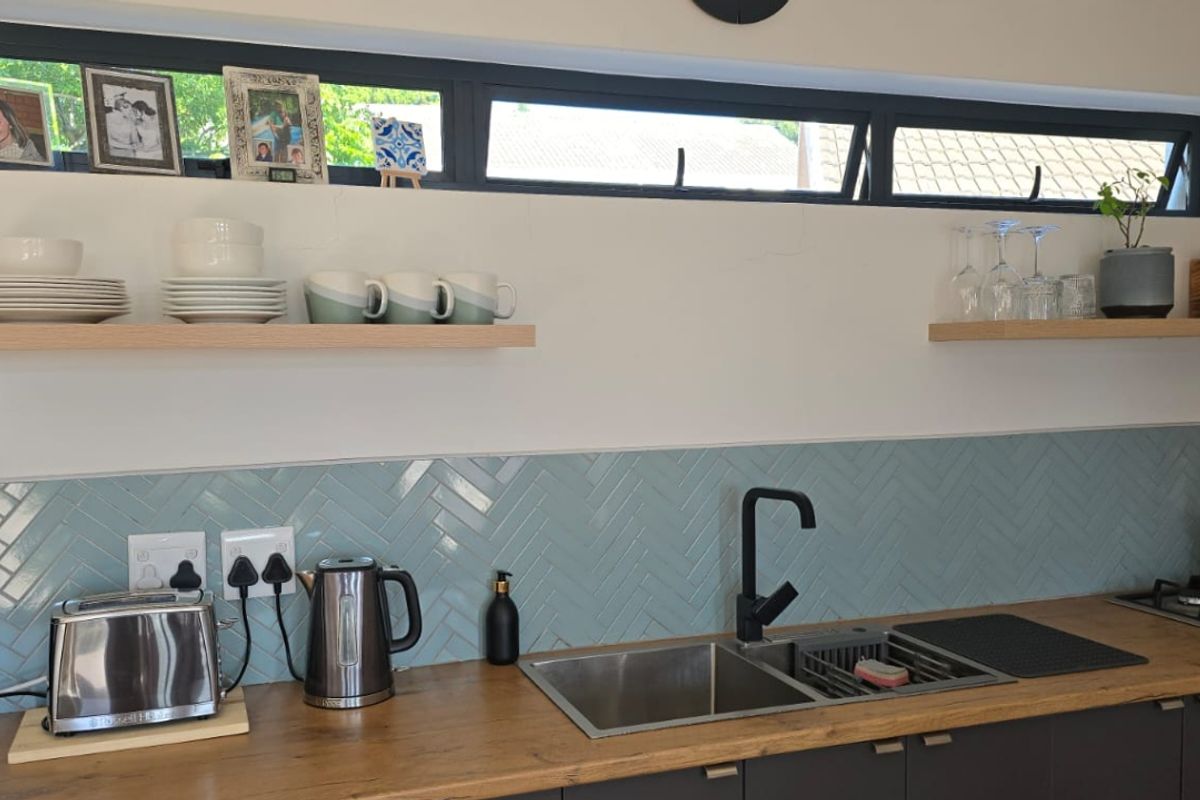 Modern kitchen featuring a stylish light blue herringbone backsplash, timber countertops, floating shelves, and integrated stainless steel sink with black fixtures.