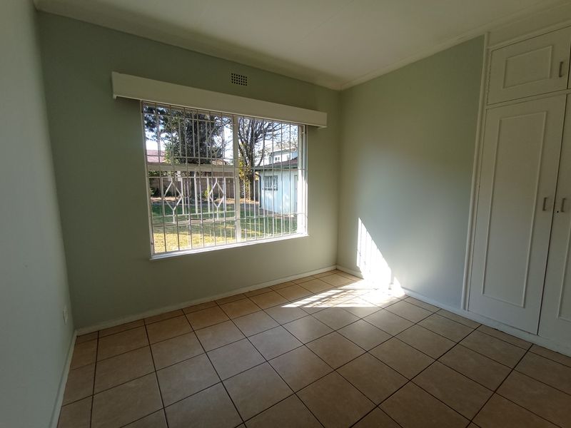 A bright, empty bedroom featuring tiled flooring, built-in cupboards, and a large window fitted with security bars.