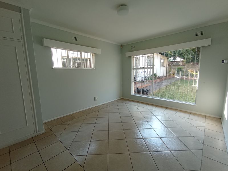 A bright, empty bedroom featuring tiled flooring, built-in cupboards, and large windows fitted with security bars overlooking the garden.