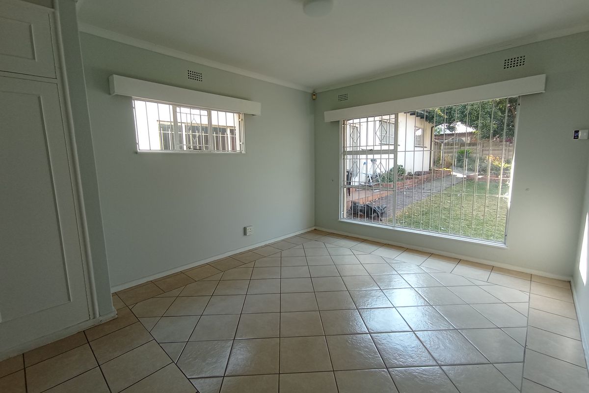 A bright, empty bedroom featuring tiled flooring, built-in cupboards, and large windows fitted with security bars overlooking the garden.