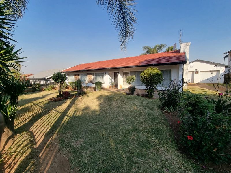 A rear view of a single-story residential house featuring a tiled roof, brick chimney, and a well-maintained lawn with established garden beds.