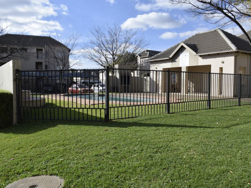 A communal swimming pool area featuring a secure metal fence, paved surround, and an adjacent covered patio structure within a well-maintained residential complex.