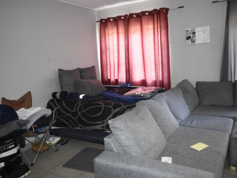 A living room area featuring tiled flooring and a window with a curtain rod, currently showing signs of wear on the wall surfaces.