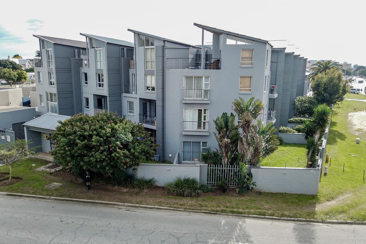 Aerial view of a modern multi-story apartment complex featuring grey facade, balconies, and landscaped garden area with perimeter fencing.