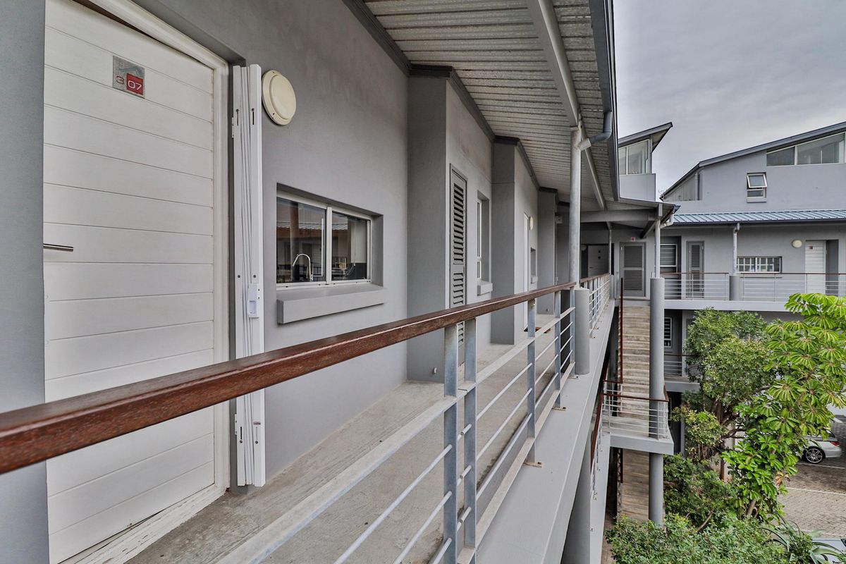Exterior view of an apartment building walkway featuring a metal railing, concrete flooring, and access doors to individual units.