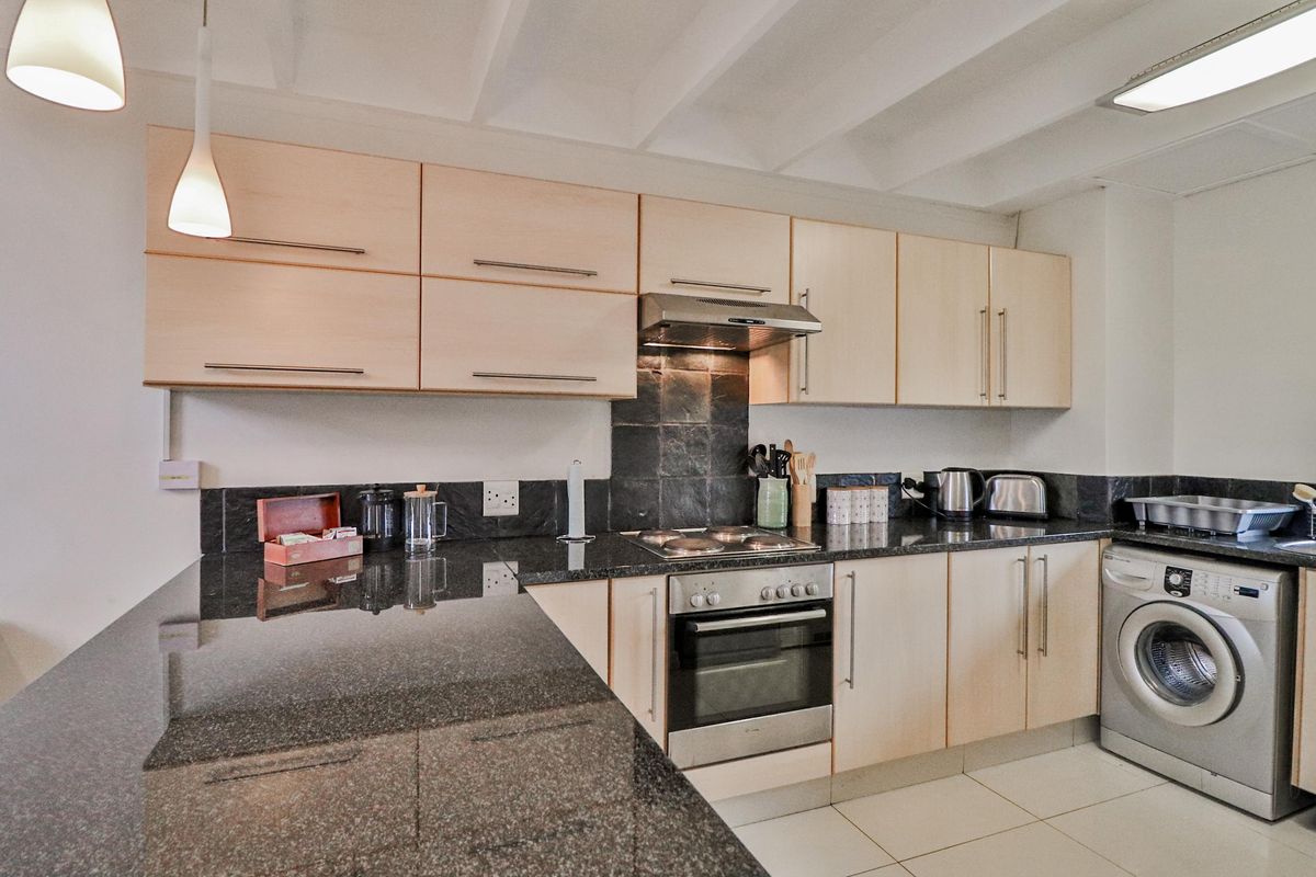 A functional kitchen featuring light wood cabinetry, dark granite countertops, a built-in oven with hob, extractor fan, and an integrated washing machine.