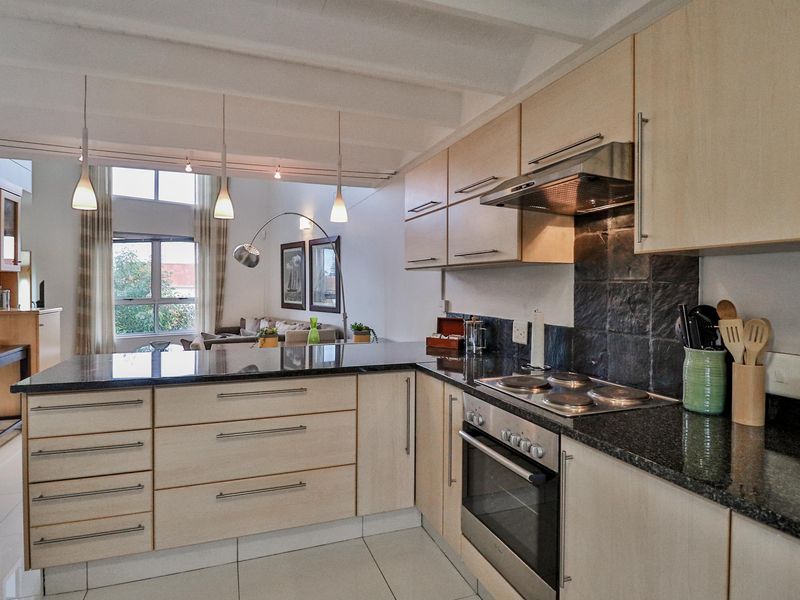 Open-plan kitchen featuring light wood cabinetry, granite countertops, built-in oven, and extractor fan, flowing into a bright living area with high ceilings.
