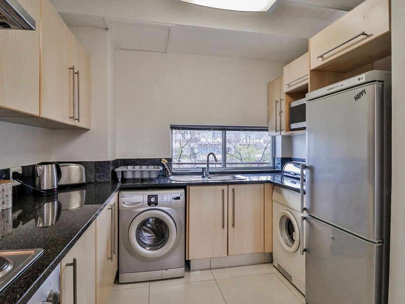 Compact kitchen featuring light wood cabinetry, dark granite countertops, stainless steel sink, and integrated laundry appliance space.