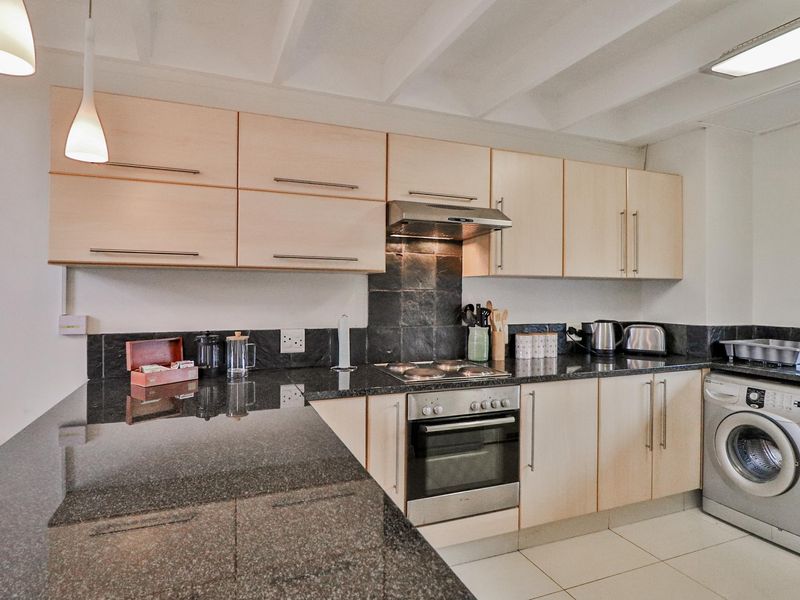 A functional kitchen featuring light wood cabinetry, dark granite countertops, a built-in oven with hob, extractor fan, and an integrated washing machine.