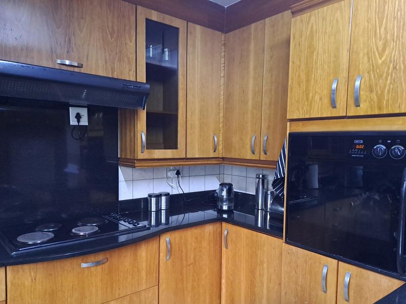 A functional kitchen featuring warm-toned wooden cabinetry, contrasting black granite countertops, a built-in oven, and an integrated electric hob with an extractor fan.