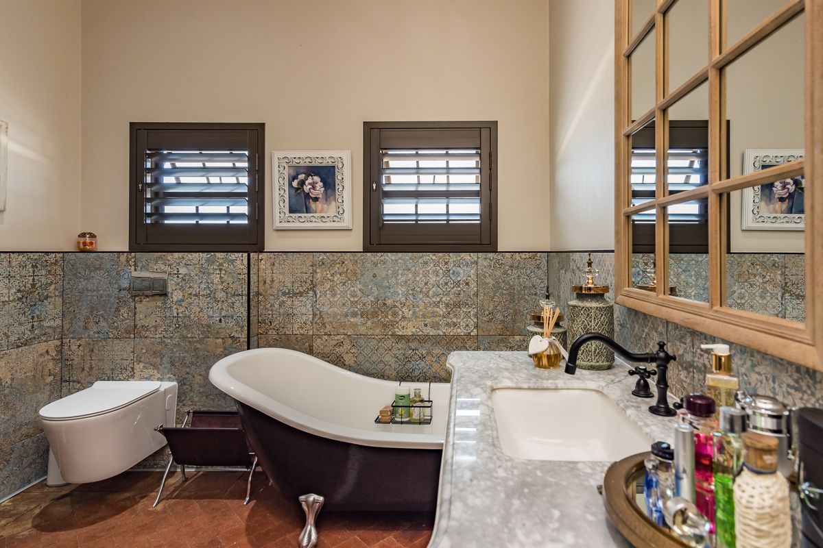 A well-appointed bathroom featuring a freestanding bathtub, wall-mounted toilet, marble-topped vanity, and decorative wall tiles with shuttered windows.
