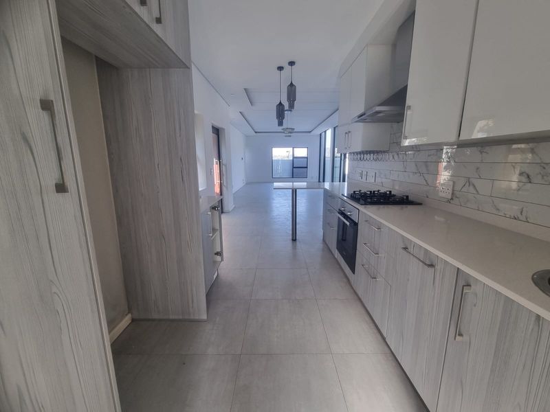 Modern open-plan kitchen featuring sleek wood-grain cabinetry, stone countertops, integrated gas hob, and oven, flowing into a spacious, light-filled living area with tiled flooring.