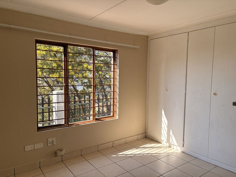 A bright, empty bedroom featuring tiled flooring, built-in cupboards, and a window with security bars overlooking trees.