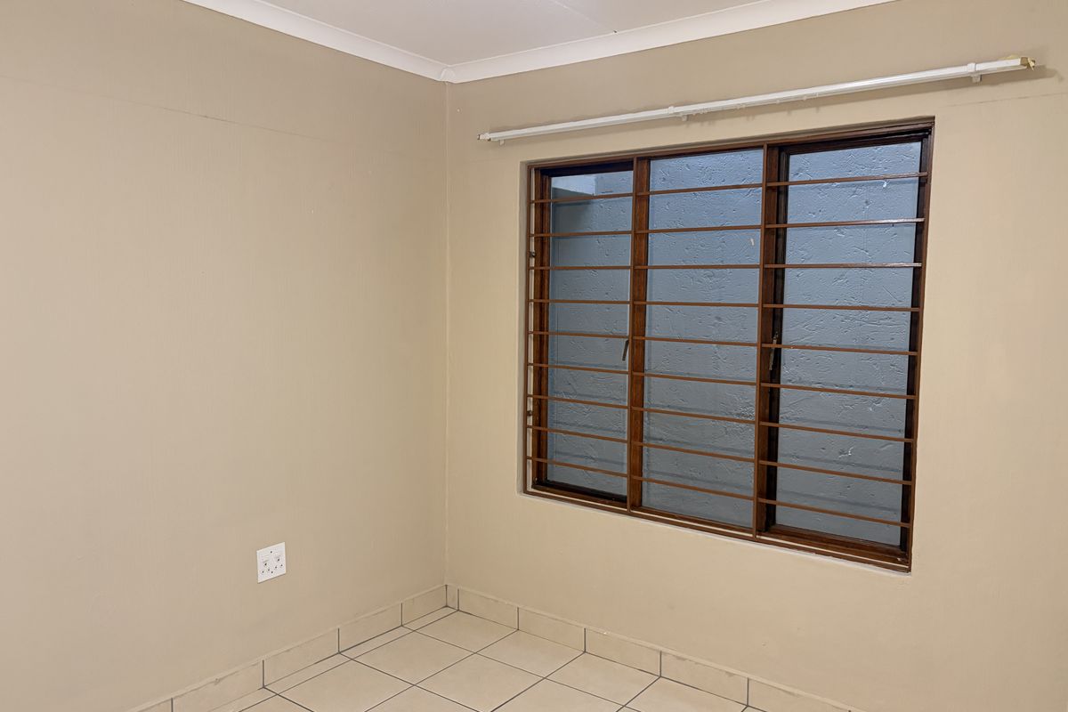 A clean, empty bedroom featuring neutral wall paint, tiled flooring, and a window fitted with security bars.