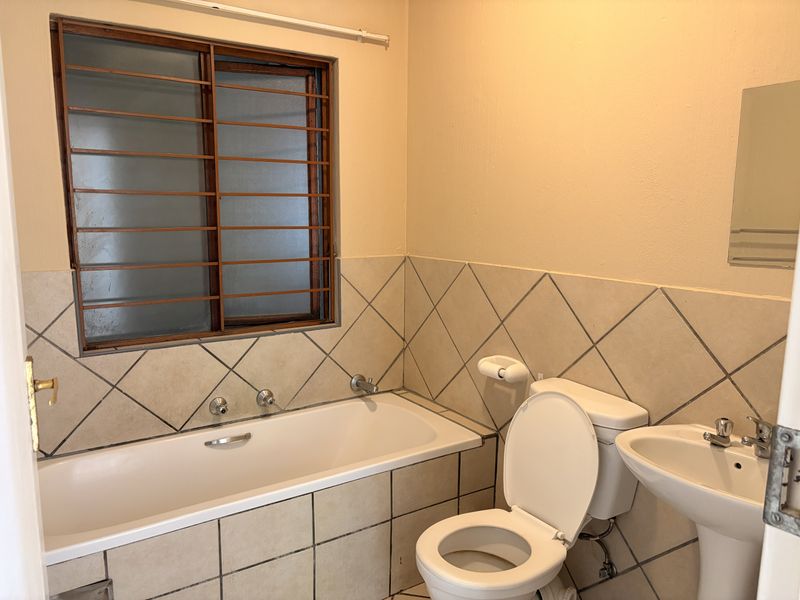 A functional bathroom featuring a bathtub, pedestal basin, and toilet, finished with neutral wall tiles and fitted with security bars on the window.