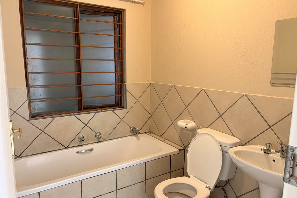 A functional bathroom featuring a bathtub, pedestal basin, and toilet, finished with neutral wall tiles and fitted with security bars on the window.