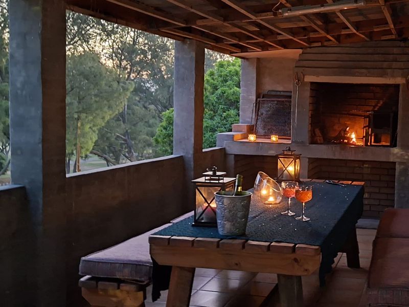 Covered outdoor patio featuring a built-in brick fireplace, exposed timber ceiling beams, and tiled flooring, overlooking a garden area.