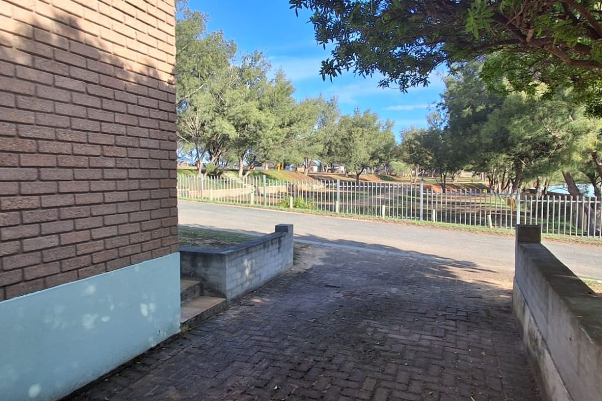 A view of the rear exterior area featuring a brick wall, paved driveway, and metal fencing overlooking a landscaped green space.