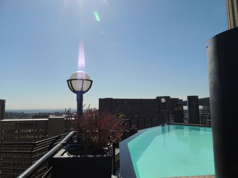 A rooftop swimming pool area featuring modern decking, metal safety railings, and panoramic city views under a clear sky.
