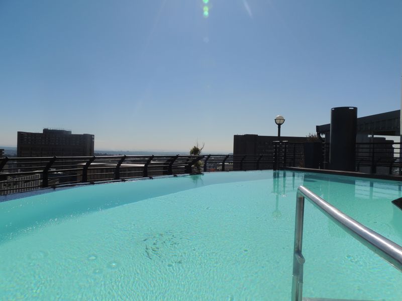 A rooftop infinity swimming pool featuring a stainless steel ladder, metal safety railings, and expansive city views under a clear sky.