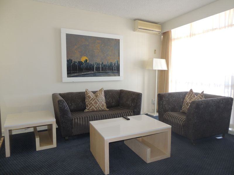 A living room area featuring a wall-mounted air conditioning unit, fitted carpeting, and large windows allowing for natural light.