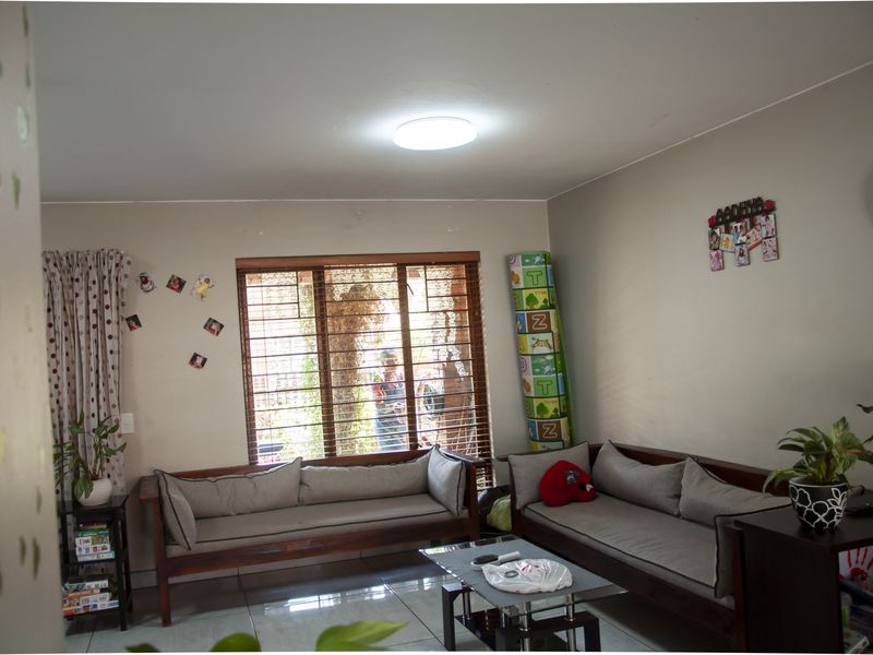 A living room featuring tiled flooring, a large window with wooden blinds, and security bars, providing a functional and well-lit living space.