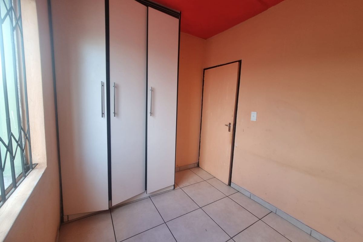 A compact bedroom featuring tiled flooring, a built-in wardrobe, and a security gate on the window. The room is currently vacant.