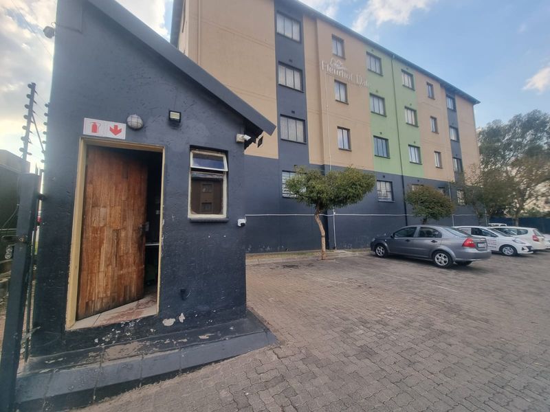 Exterior view of the apartment complex entrance featuring a security guard house, paved parking area, and perimeter electric fencing.
