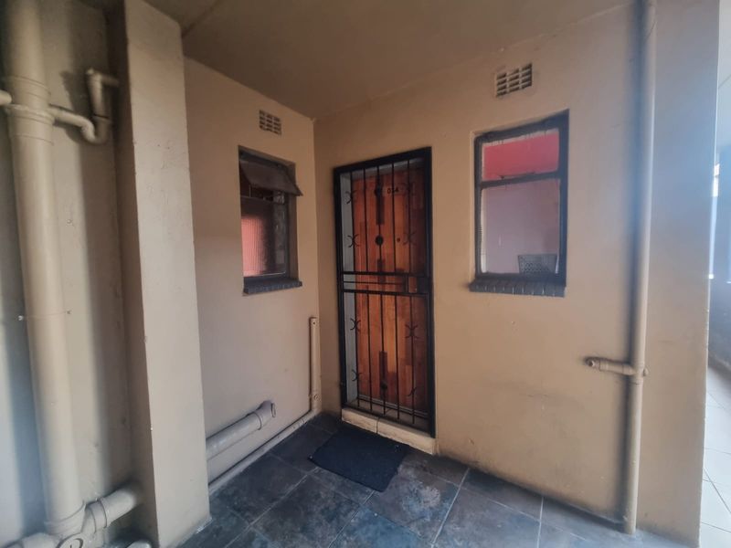 Apartment entrance area featuring a security gate, timber door, and tiled flooring with visible external plumbing and ventilation grilles.