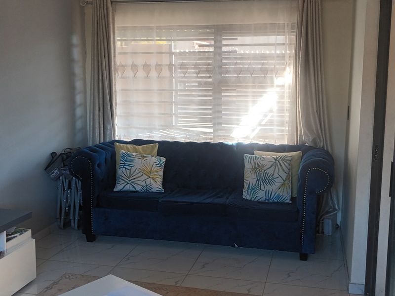 A comfortable living room featuring tiled flooring and large windows with venetian blinds and curtains, providing natural light to the space.