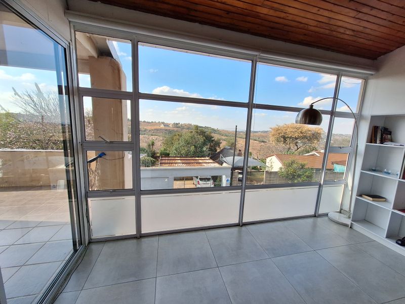 Bright living area featuring expansive floor-to-ceiling windows with scenic views, tiled flooring, and a warm timber-clad ceiling, offering a versatile and light-filled space.