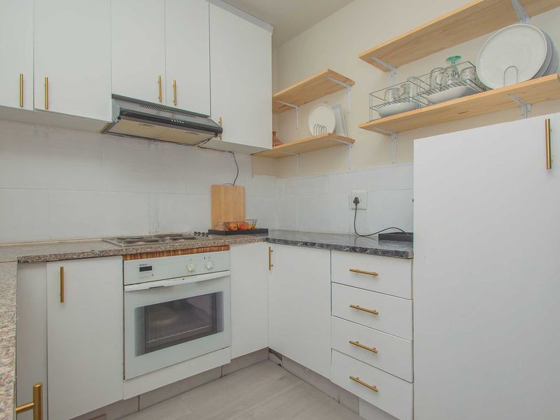 A compact, modern kitchen featuring white cabinetry, granite countertops, built-in oven, electric hob, and open wooden shelving for storage.