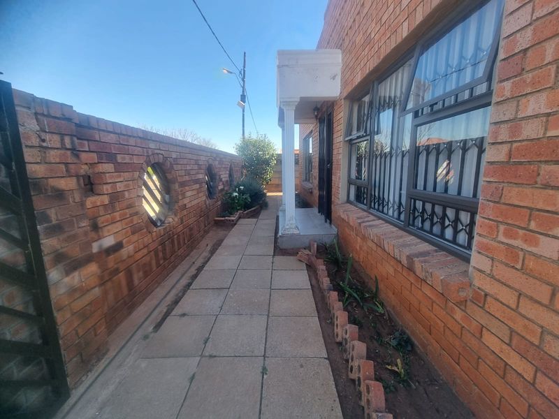 A side entrance pathway featuring face brick walls, security bars on windows, and a paved walkway leading to a covered porch area.