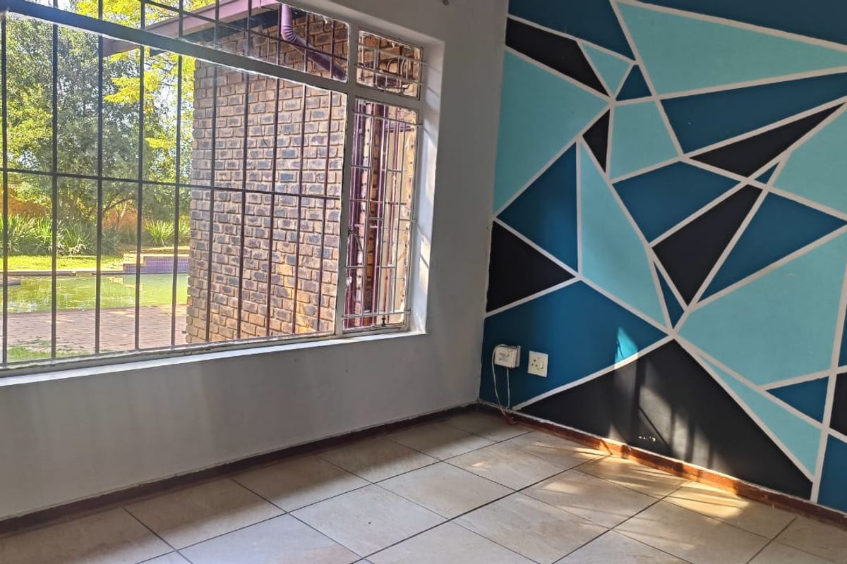 A bright living area featuring tiled flooring, a geometric accent wall, and large windows with security bars overlooking the garden and pool area.