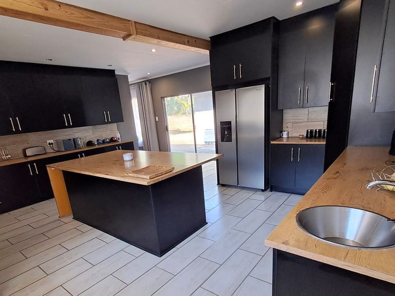 Modern kitchen featuring dark cabinetry, a central island with a wooden countertop, tiled flooring, and exposed timber ceiling beams with access to the outdoors.