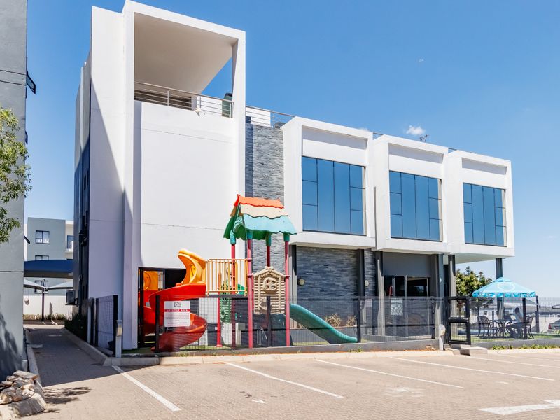 Modern apartment building exterior featuring stone cladding, large glass windows, private balconies, and a paved parking area with an adjacent children's play area.