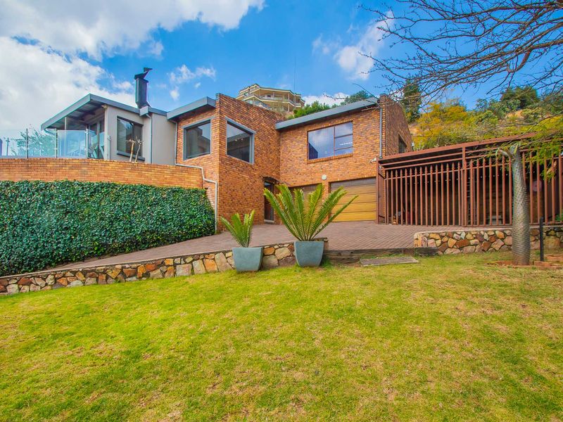 Rear view of a multi-level brick house featuring a double garage, elevated terrace with glass balustrade, and landscaped garden area.