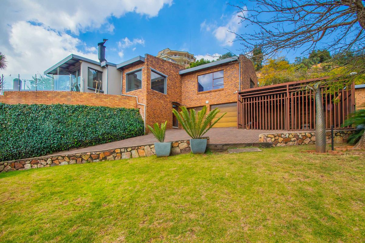 Rear view of a multi-level brick house featuring a double garage, elevated terrace with glass balustrade, and landscaped garden area.