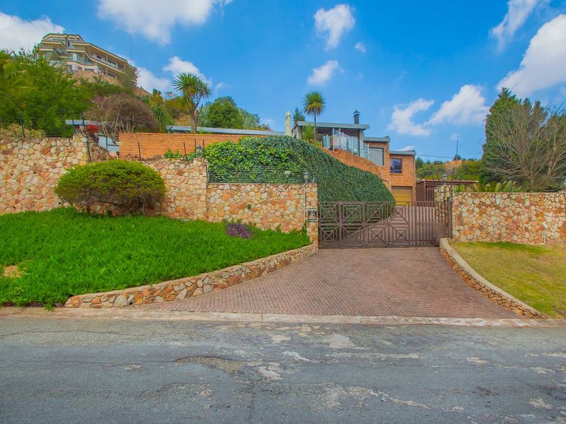 Front view of the property featuring a paved driveway, secure metal gate, and extensive stone retaining walls set against a landscaped hillside.