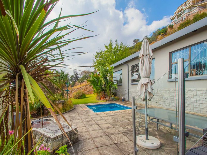 Outdoor entertainment area featuring a paved patio, built-in braai, and a swimming pool, set against a landscaped garden backdrop.