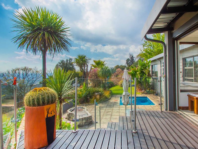An elevated timber deck featuring a glass balustrade, overlooking a paved patio area with a swimming pool and stone fire pit.