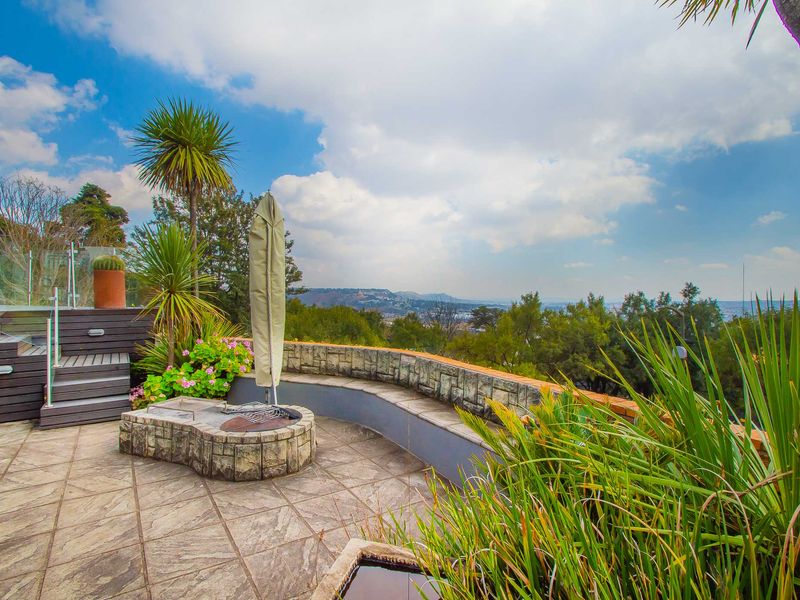 An elevated outdoor patio featuring a stone-clad built-in braai, timber decking, and a glass balustrade, offering expansive views of the surrounding landscape.