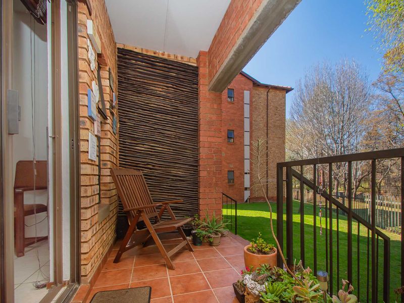 A covered balcony featuring exposed brick walls, tiled flooring, and a metal balustrade overlooking a communal garden area.