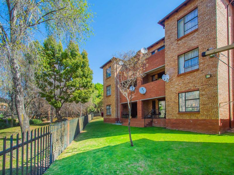 Rear view of a multi-story face brick apartment building featuring balconies, steel-framed windows, and a well-maintained communal garden area with perimeter fencing.