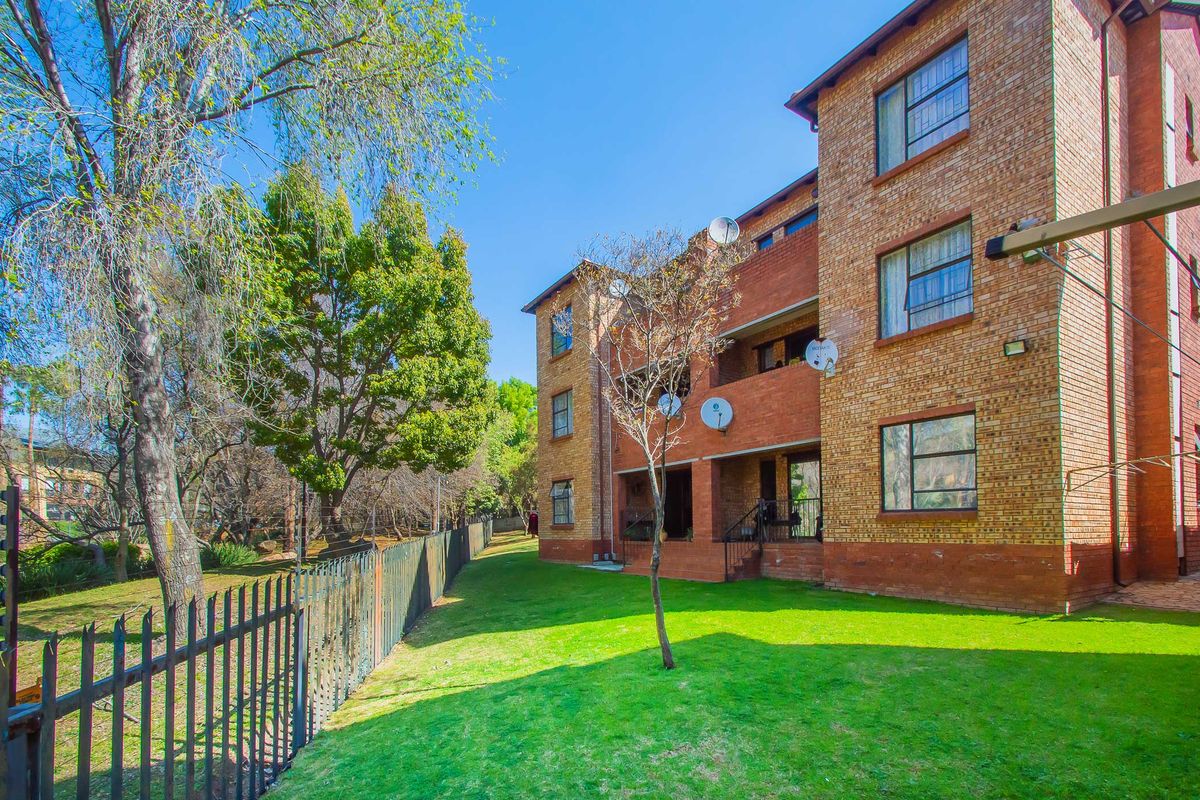 Rear view of a multi-story face brick apartment building featuring balconies, steel-framed windows, and a well-maintained communal garden area with perimeter fencing.