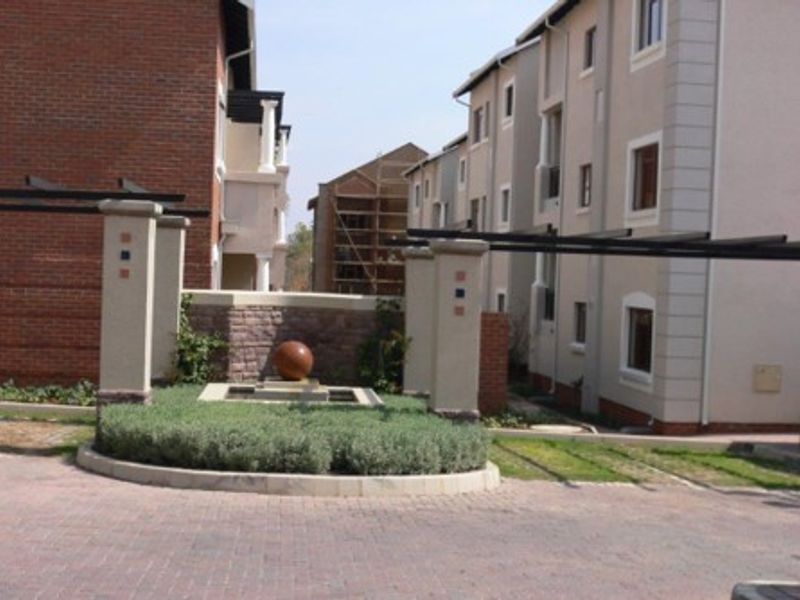 Front entrance view of an apartment complex featuring a paved driveway, landscaped garden bed, and a central water feature.